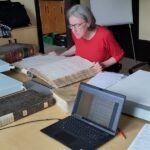 Photographie en couleurs d'un ordinateur posé sur une table et d'un être humain qui consulte de gros registres d'archives
